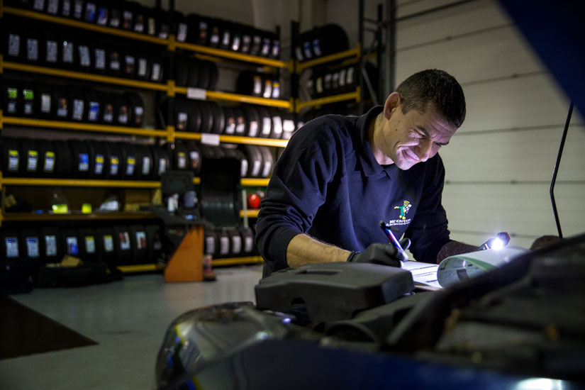 Mechanic holding a clipboard leaning over conducting an MOT test in a garage