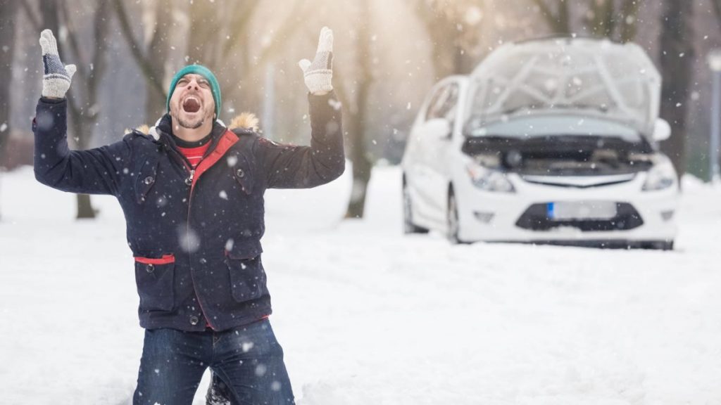 Man on his knees with arms raised screaming in frustration while their car has broken-down behind in a snowy condition