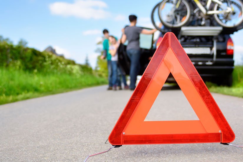 Red warning triangle placed on a road in front of a family loading their car for a summer road trip
