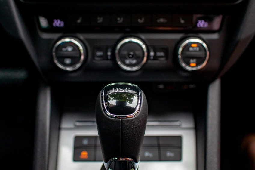 Close-up of a DSG gear lever inside a car, with control dials and the dashboard softly blurred in the background.